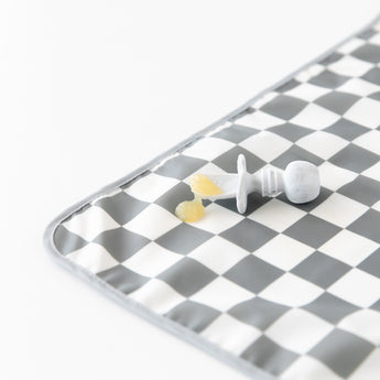 A Bumkins Silicone Chewtensils®: Marble lays atop a gray & white checkered mat on a white background with applesauce on top.