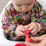 A baby wearing a Super Mario™ Mashup sleeved bib, touches a Bumkins Silicone Chewtensils®: Red with cottage cheese on it.