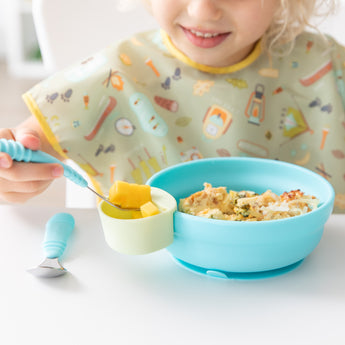 Child eating with Bumkins Spoon + Fork: Blue. Rice, veggies in a bowl, and mango slice in a cup. Colorful bib worn.