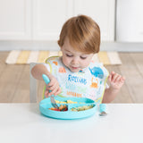 Toddler uses Bumkins Blue Spoon + Fork to eat from a blue plate, wearing Rolling with the waves bib at a white table.