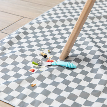 A Bumkins Spoon + Fork: Blue lays atop a gray & white checkered floor mat among scattered bits of fruit & veggies by a high chair leg.