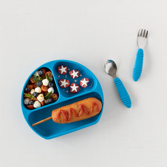 Blue tray with fruit salad, star snacks, corndog; Bumkins Spoon + Fork: Dark Blue for little hands beside it.