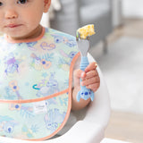 Close-up of a toddler in a pastel bib holding a Bumkins Spoon + Fork: Stitch with scrambled eggs.