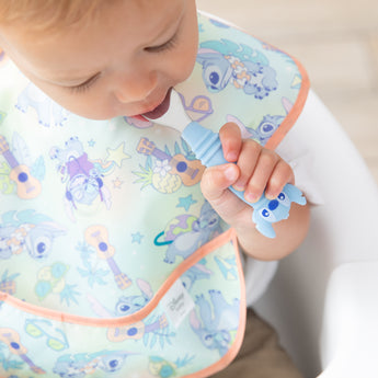Close-up of a toddler wearing a pastel bib in a high chair with a Bumkins Spoon + Fork: Stitch in their mouth.