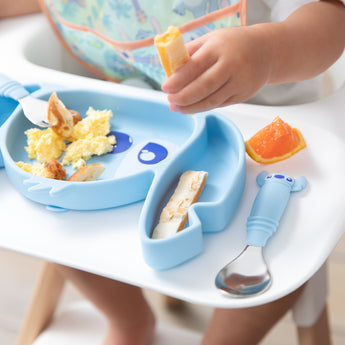 A Bumkins Spoon + Fork: Stitch sits on a high chair tray next to a Stitch-shaped dish with breakfast foods; a child’s hand visible above.