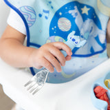 Close-up of a child in a blue bib seated in a high chair, holding a fork from Bumkins Spoon + Fork: Stitch.