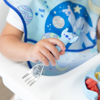 Close-up of a child in a blue bib seated in a high chair, holding a fork from Bumkins Spoon + Fork: Stitch.