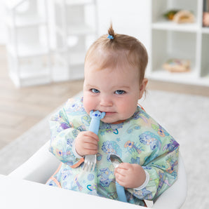 A baby with a topknot smiles in a high chair, holding Bumkins Spoon + Fork: Stitch in a bright playroom.