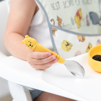 Close-up of a child's hand holding a Bumkins Spoon + Fork: Winnie the Pooh on a high chair tray.