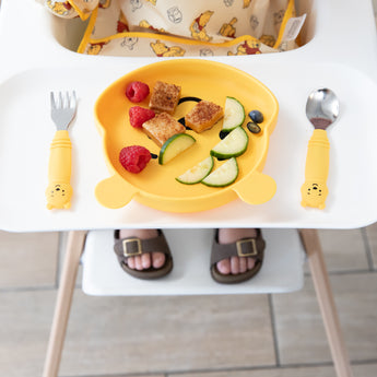 Bumkins Spoon + Fork: Winnie the Pooh sits beside a matching plate with fruit & veggies on a high chair tray; a child's feet visible below.