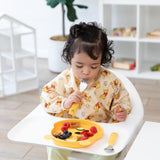 A child in a yellow patterned bib sits in a high chair, holding a Bumkins Spoon + Fork: Winnie the Pooh near a bear-shaped plate of food.