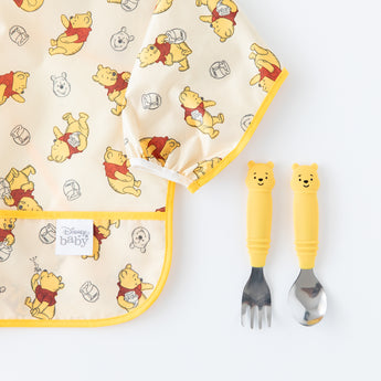 Bumkins Spoon + Fork: Winnie the Pooh sits next to a yellow Winnie the Pooh patterned bib on a white background.