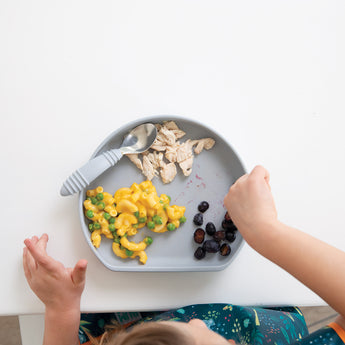 A child enjoys shredded chicken, mac and cheese with peas, and grapes using a Bumkins Spoon + Fork: Gray from their utensil set.