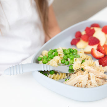 A child enjoys a divided plate of pasta, peas, and apples with their Bumkins Spoon + Fork: Gray set—ideal for self-feeding.