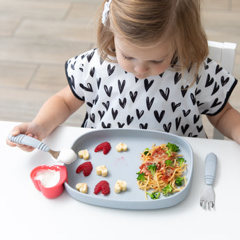 A young child enjoys spaghetti, cheese, and raspberries using Bumkins Spoon + Fork: Gray on a gray plate in a white high chair.