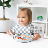 A toddler in a checkered bib smiles, using Bumkins Spoon + Fork: Gray, while holding a divided plate with various foods.