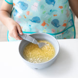 Someone in a colorful apron mixes food in a light blue bowl with a Bumkins Spoon + Fork: Gray on a white table.
