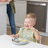 A child joyfully self-feeds with Bumkins Spoon + Fork in gray, using a sectioned silicone plate as an adult watches close by.
