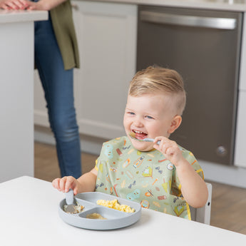 A child joyfully self-feeds with Bumkins Spoon + Fork in gray, using a sectioned silicone plate as an adult watches close by.