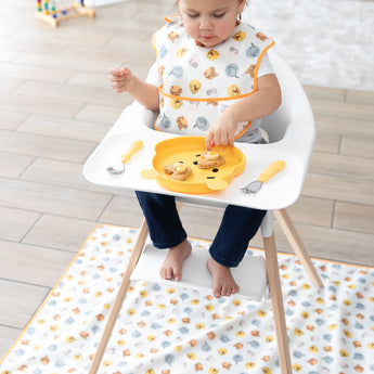 A toddler eats snacks with Bumkins Spoon + Fork: Pineapple from a yellow bear plate, wearing a bib on an animal-print mat.