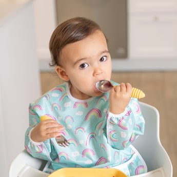 A toddler in a rainbow bib sits in a high chair using Bumkins Spoon + Fork: Pineapple, looking up at the camera.