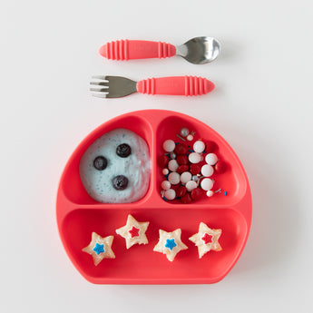 Red divided plate with yogurt, blueberries, and star snacks. Bumkins Spoon + Fork: Red above. Food-safe silicone for self-feeding!.