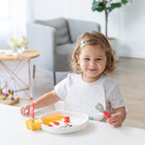A smiling toddler uses Bumkins Spoon + Fork in Red, self-feeding with sliced fruits at a table with a bib.