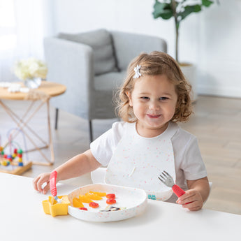 A smiling toddler uses Bumkins Spoon + Fork in Red, self-feeding with sliced fruits at a table with a bib.