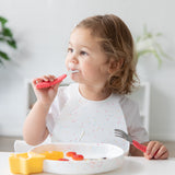 A toddler with light brown hair smiles while self-feeding at a table. Theyre using a Bumkins red silicone spoon, wearing a white speckled bib, and eating fruit from a white dish. A yellow star-shaped cup is close by.