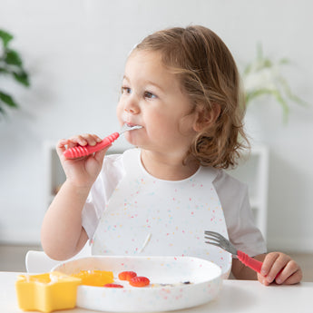 A toddler with light brown hair smiles while self-feeding at a table. Theyre using a Bumkins red silicone spoon, wearing a white speckled bib, and eating fruit from a white dish. A yellow star-shaped cup is close by.