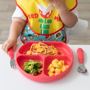 A toddler uses Bumkins red Spoon + Fork to self-feed spaghetti, broccoli, and pineapple from a red divided plate with a colorful bib.