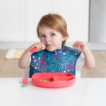 A toddler uses Bumkins Spoon + Fork: Red to eat from a red plate at a white table, wearing a colorful bib.