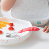 A child uses Bumkins toddler utensils, holding a Spoon + Fork: Red with a white speckled plate of sliced strawberries and orange.