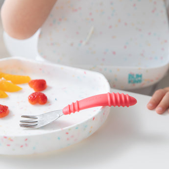 A child uses Bumkins toddler utensils, holding a Spoon + Fork: Red with a white speckled plate of sliced strawberries and orange.