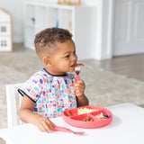 A child self-feeds with Spoon + Fork: Red, using a colorful divided plate and wearing a patterned bib from Bumkins at the table.
