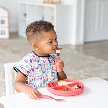 A child self-feeds with Spoon + Fork: Red, using a colorful divided plate and wearing a patterned bib from Bumkins at the table.