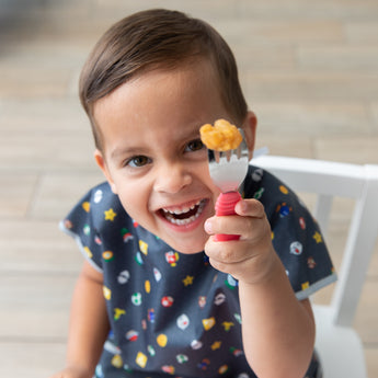 A smily boy in a Bumkins Junior Bib: Super Mario™ Lineup sits happily in a white chair holding Red Spoon + Fork Set in front of his face.