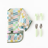 Bumkins Sleeved Bib: Motley Check lays folded next to two sets of light green toddler utensils on a white background.