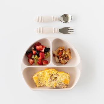 A divided plate with an omelette, fruit, and potatoes sits beneath Bumkins Spoon + Fork: Sand on a white background.