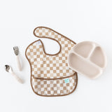 Bumkins Spoon + Fork: Sand sits next to the left of a tan checkered bib and a beige sectioned plate on a white background.