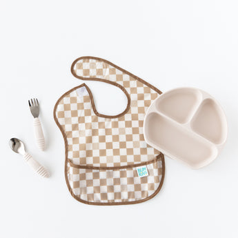 Bumkins Spoon + Fork: Sand sits next to the left of a tan checkered bib and a beige sectioned plate on a white background.