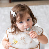 A girl wearing a beige bib holds Bumkins Spoon + Fork: Sand, one in her mouth, as she looks curiously at the camera.