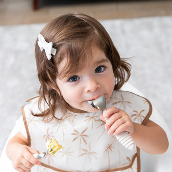 A girl wearing a beige bib holds Bumkins Spoon + Fork: Sand, one in her mouth, as she looks curiously at the camera.