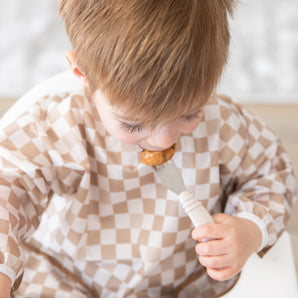 A boy in a tan checkered bib uses Bumkins Spoon + Fork: Sand to eat a meal in a high chair.