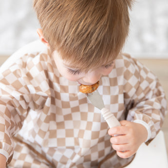 A boy in a tan checkered bib uses Bumkins Spoon + Fork: Sand to eat a meal in a high chair.