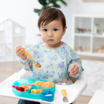 Child in a pastel bib holding a Bumkins Spoon + Fork: Tangerine.