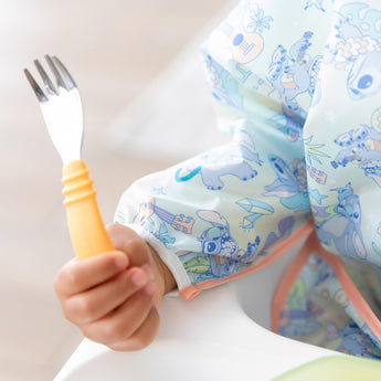 Close-up of a child in a pastel bib holding a Bumkins Spoon + Fork: Tangerine.