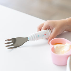 Close-up of a child's hand gripping a Bumkins Spoon + Fork: Vanilla Sprinkle at a white table next to a pink heart-shaped cup of yogurt.