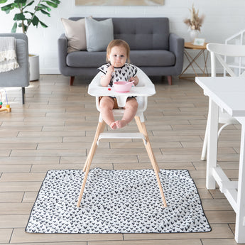 A baby in a high chair sits on a Bumkins Splat Mat: Hearts, eating from a bowl. A gray sofa and decor adorn the modern living room behind.