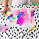 A child paints with brushes on paper at a table covered with a Bumkins Splat Mat: Hearts, with a paint palette nearby.